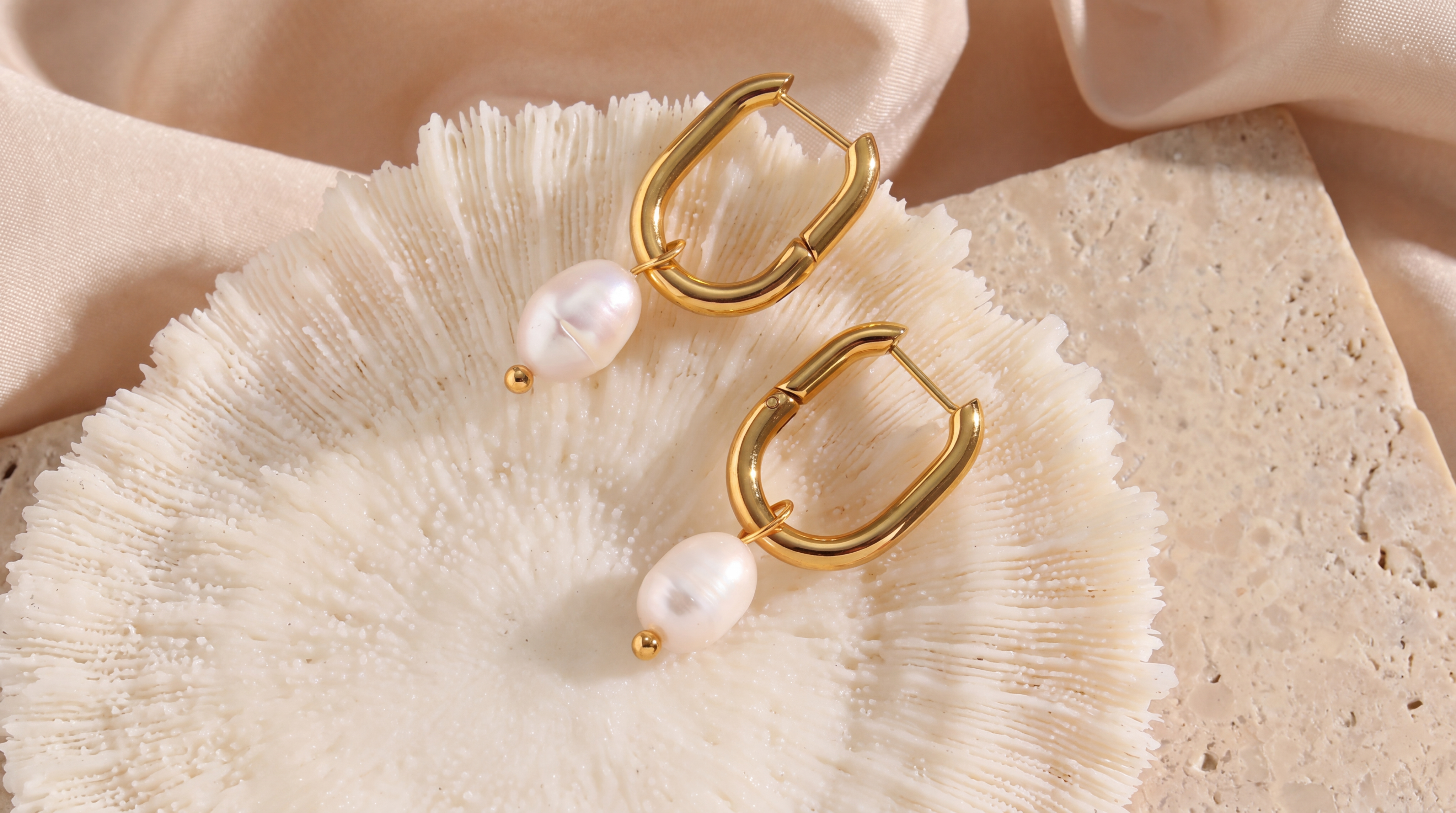 Gold hoop earrings with pearl drops resting on coral and travertine