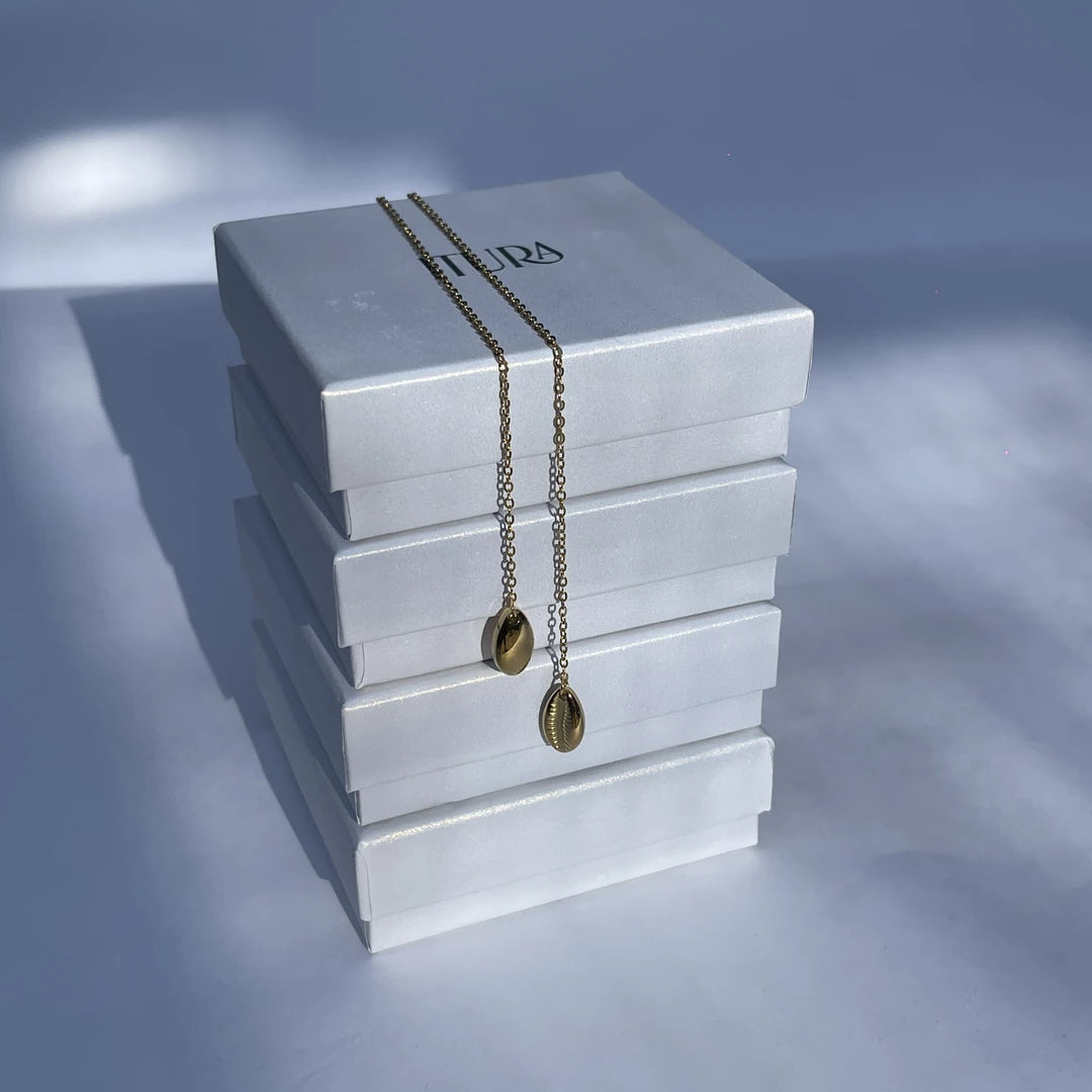 A gold chain necklace with two cowrie gold pendants is draped across a stack of white rectangular jewelry boxes against a soft blue-gray background.