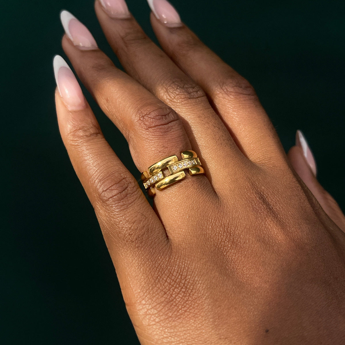 A close-up of a hand with long, manicured pink nails wearing a gold ring with a textured, interlocking chain-link design and small clear stones, set against a dark green background.